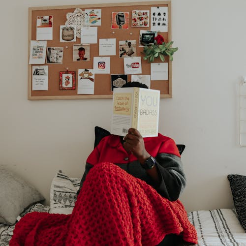 Free Person in Black and Red Jacket Sitting on Bed Reading a Book Stock Photo