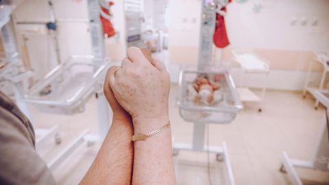 A parent's hands clasped together in a hospital nursery, with newborns in the background.