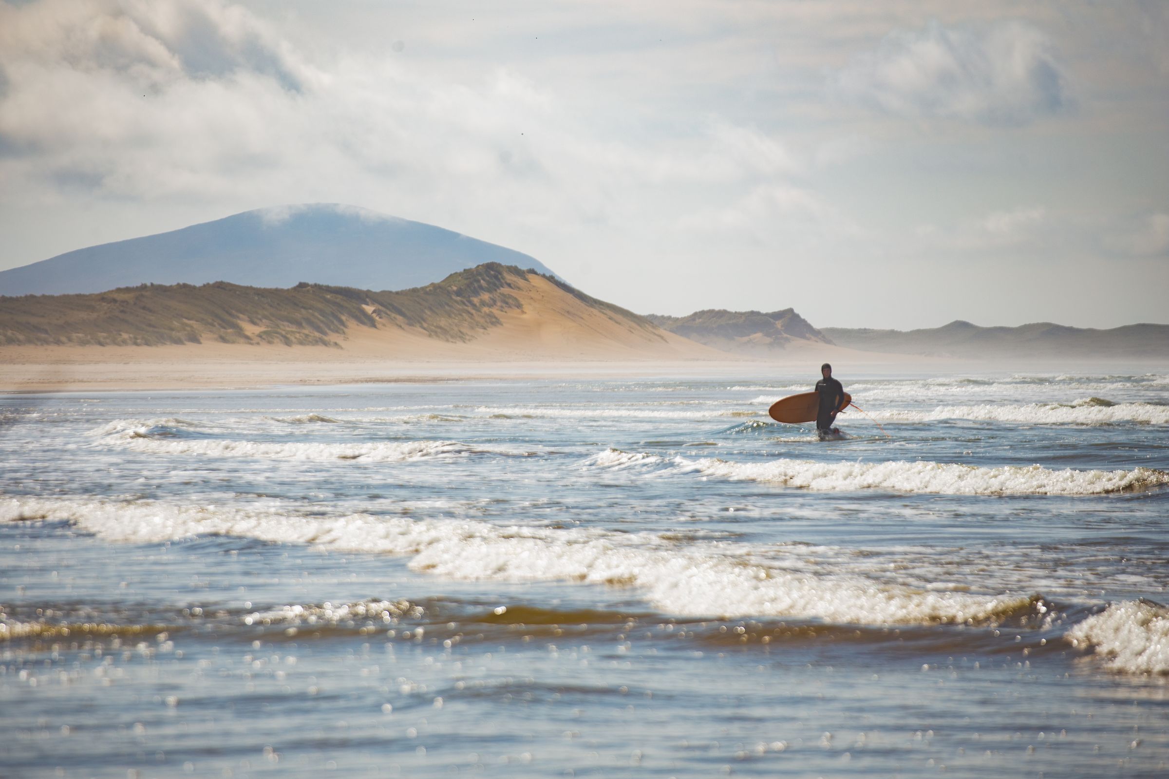 A lone surfer walks into the waves of a beach in Donegal, Ireland, with rolling hills in the background