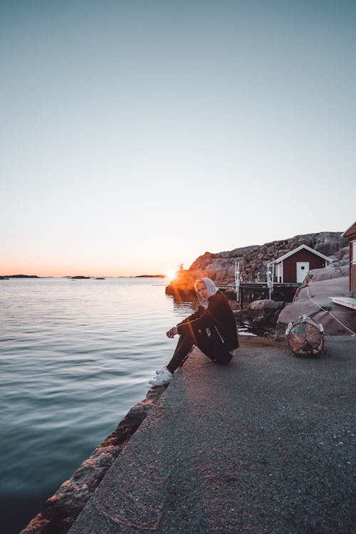 Side view of anonymous guy in casual wear resting on cement platform near rippled ocean and mounts in bright sunlight in evening in back lit under serene sky