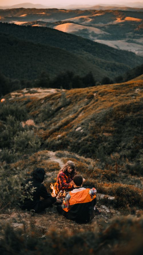 Free Three friends enjoy a campfire in the scenic mountains during sunset. Perfect for outdoor adventure themes. Stock Photo