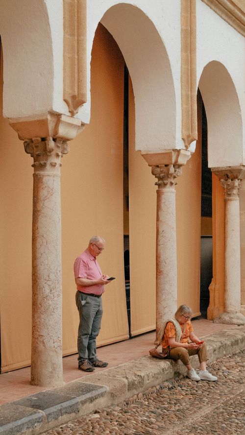 Free An elderly couple enjoys a quiet moment reading at a historic courtyard with stone columns. Stock Photo