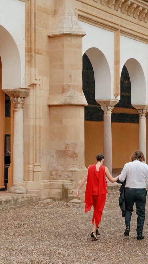 Free A couple walking hand-in-hand by a historic arched building with a classic architecture style. Stock Photo