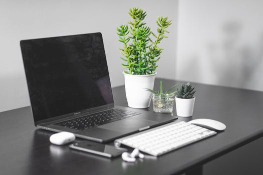 Elegant workspace featuring a MacBook, white keyboard, wireless mouse, and green plants on a minimalist black desk.