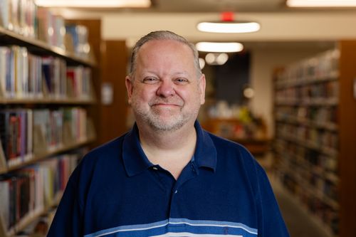 Free Portrait of a smiling man standing between bookshelves in a library. Stock Photo