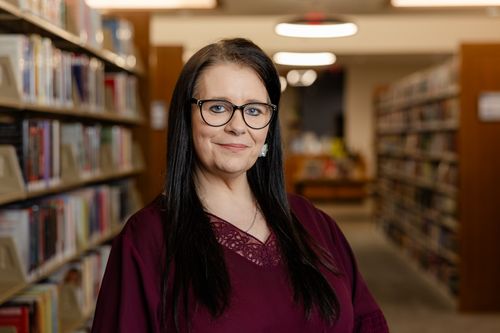 Free Confident librarian standing in a library aisle, surrounded by bookshelves. Stock Photo
