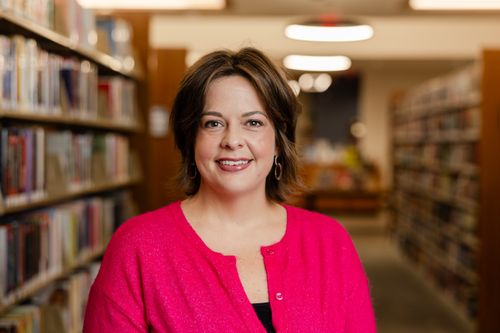 Free A confident woman in a bright pink sweater smiling in a library setting. Stock Photo