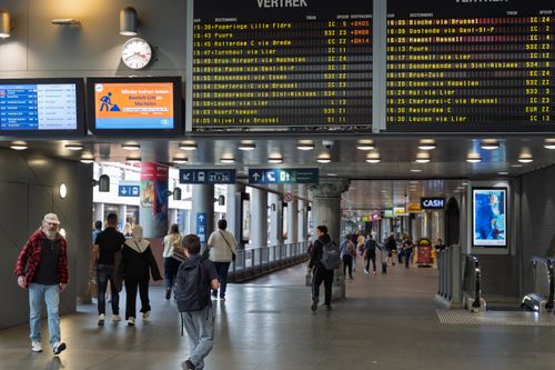 Free Commuters walking through a bustling train station under digital departure boards. Stock Photo