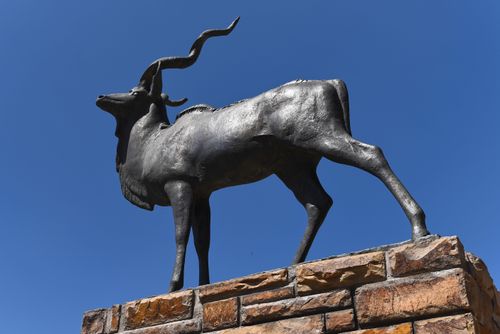 Free Bronze kudu sculpture standing on a stone base under a clear blue sky. Stock Photo