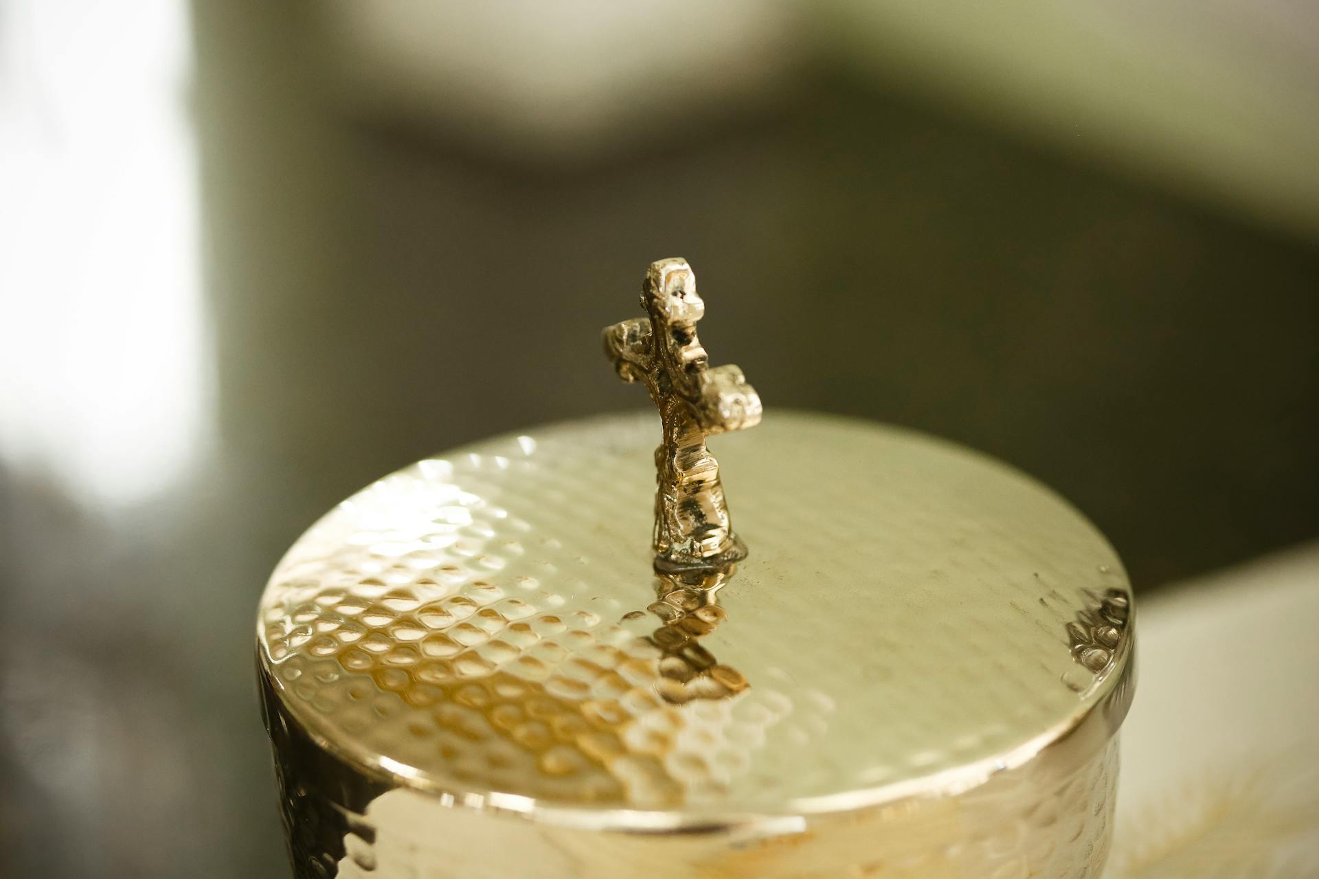 Close-up of a golden container with a textured lid and a miniature cross on top.