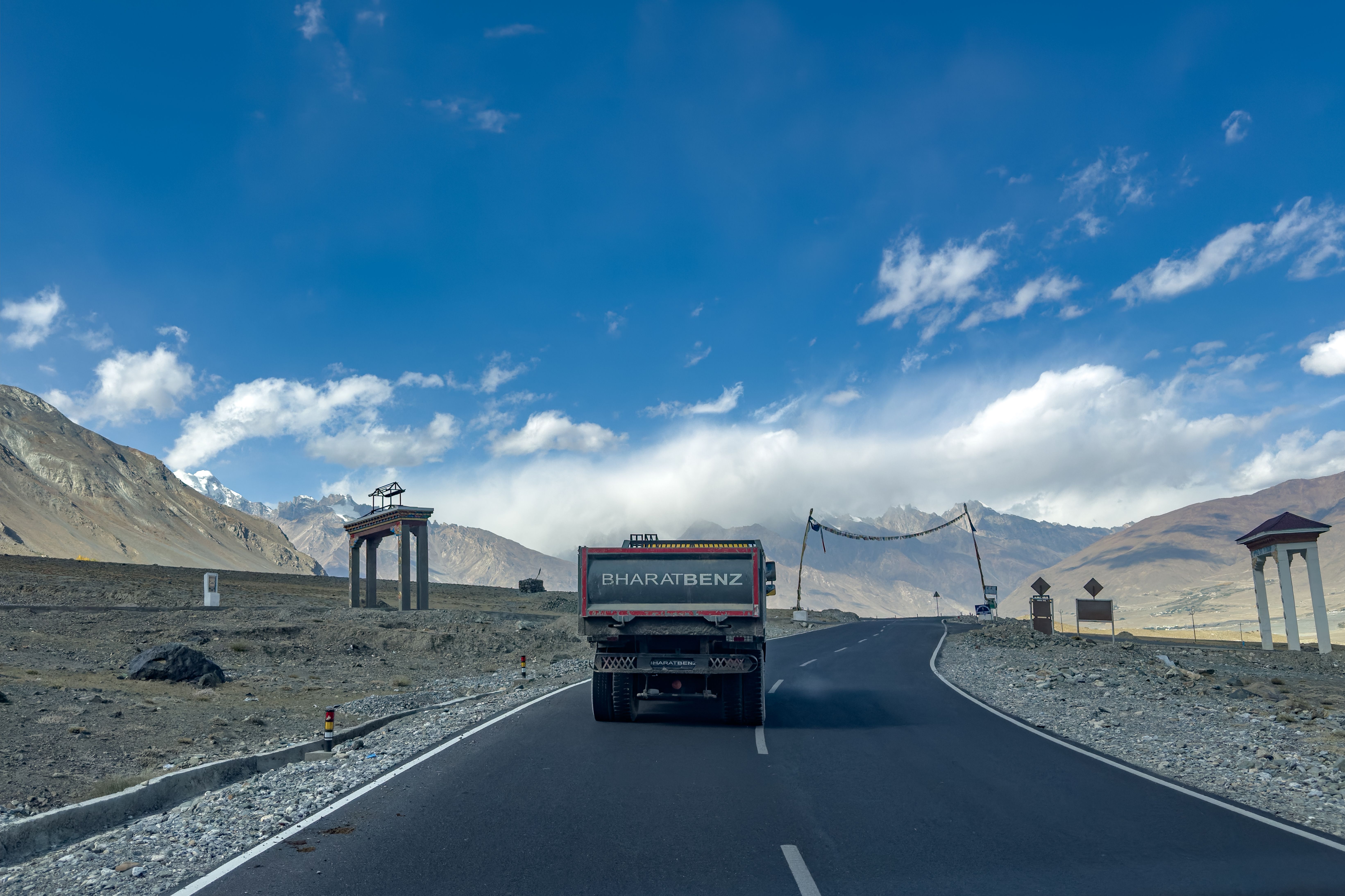 A BharatBenz truck travels on a scenic mountainous road under a clear blue sky.