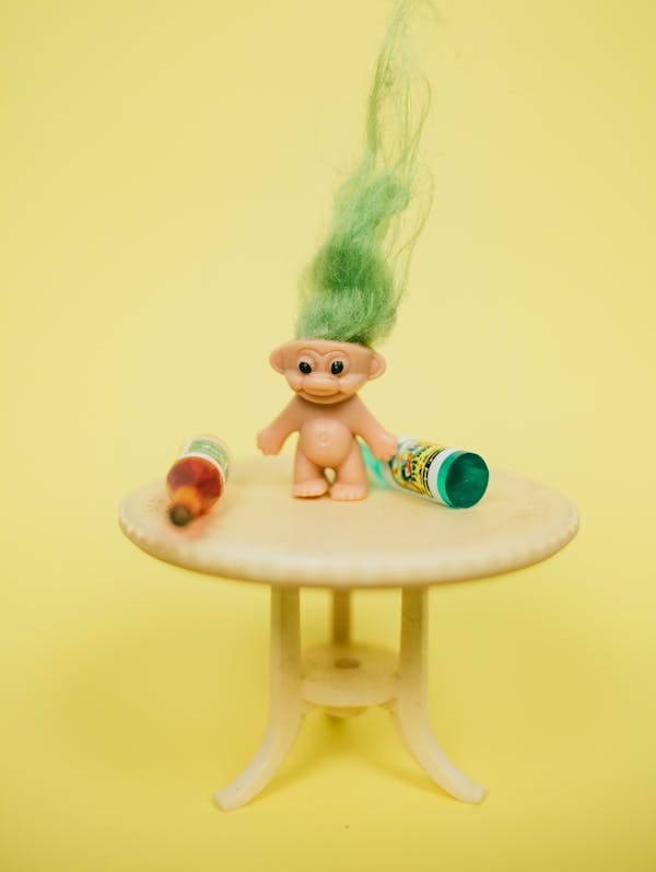 Funny troll toy with green hair placed on table miniature with various bottles against yellow background