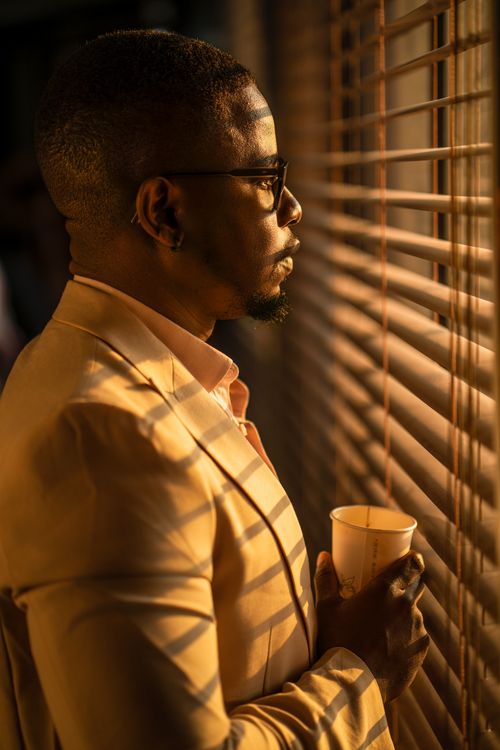 Free Profile of a man in a golden suit holding a coffee cup, bathed in warm sunlight through blinds. Stock Photo