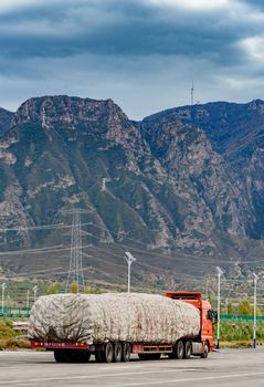 A red truck travels on a highway with a covered load against a backdrop of towering mountains and wind turbines.