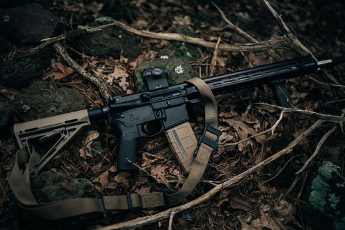 Free A tactical rifle with a sling rests on the forest floor, surrounded by leaves and branches. Stock Photo