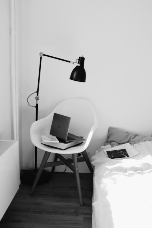 Black and white modern bedroom with laptop placed on book on white armchair under lamp near bed in light modern apartment