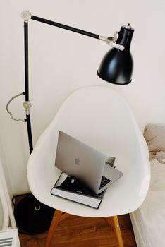 From above of portable laptop and art book composed on white armchair under lamp near wall in modern light room in apartment