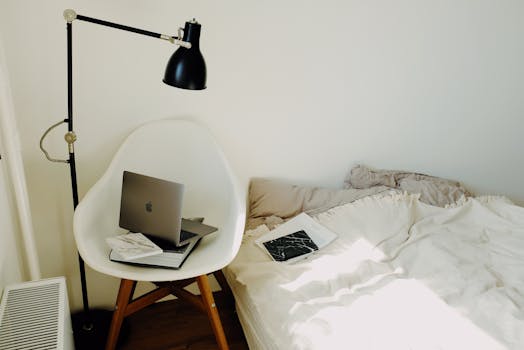 High angle of laptop placed on big black book on white armchair under lamp near comfortable bed in modern light bedroom at home