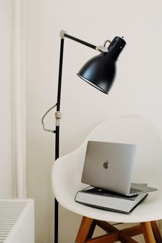 Modern workplace with laptop placed on big black book on white wooden chair under lamp near wall in light modern apartment