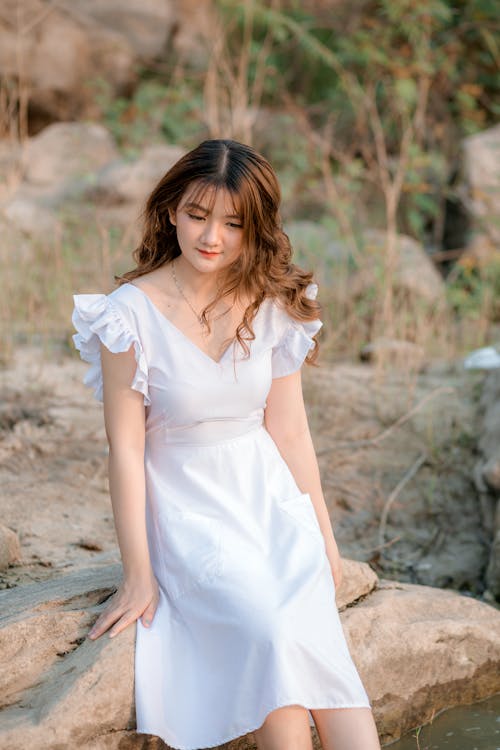 Pensive female with curly hair wearing white dress sitting on stone in daytime