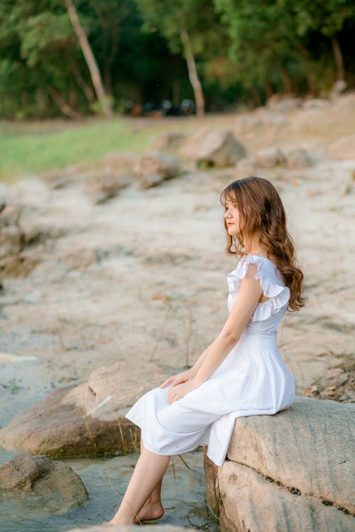 Side view of young barefooted female wearing white dress sitting on edge of stone and enjoying peaceful pond