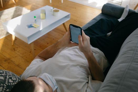 Man lying on a couch using smartphone with face mask and medication nearby, indoors.