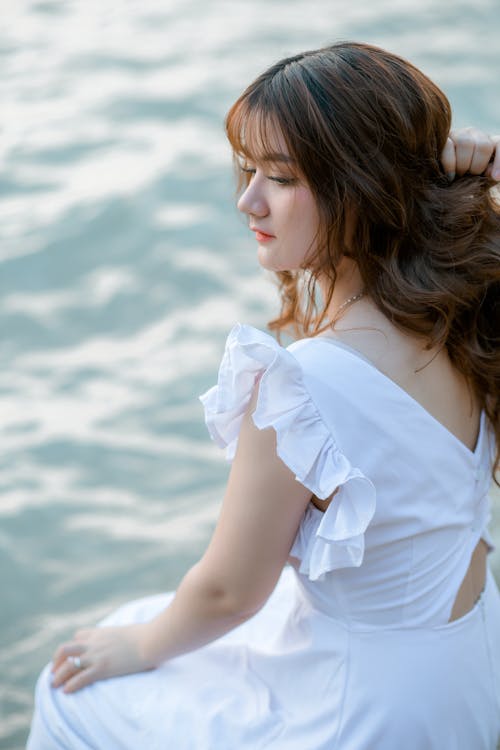 Free High angle back view of graceful young female touching hair while sitting near rippling water of lake Stock Photo