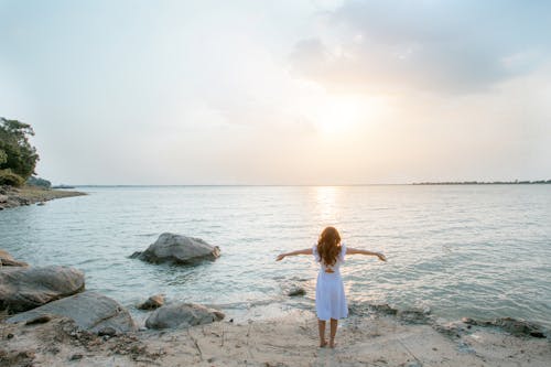 Free Woman in White Shirt Standing on Gray Rock Near Body of Water Stock Photo
