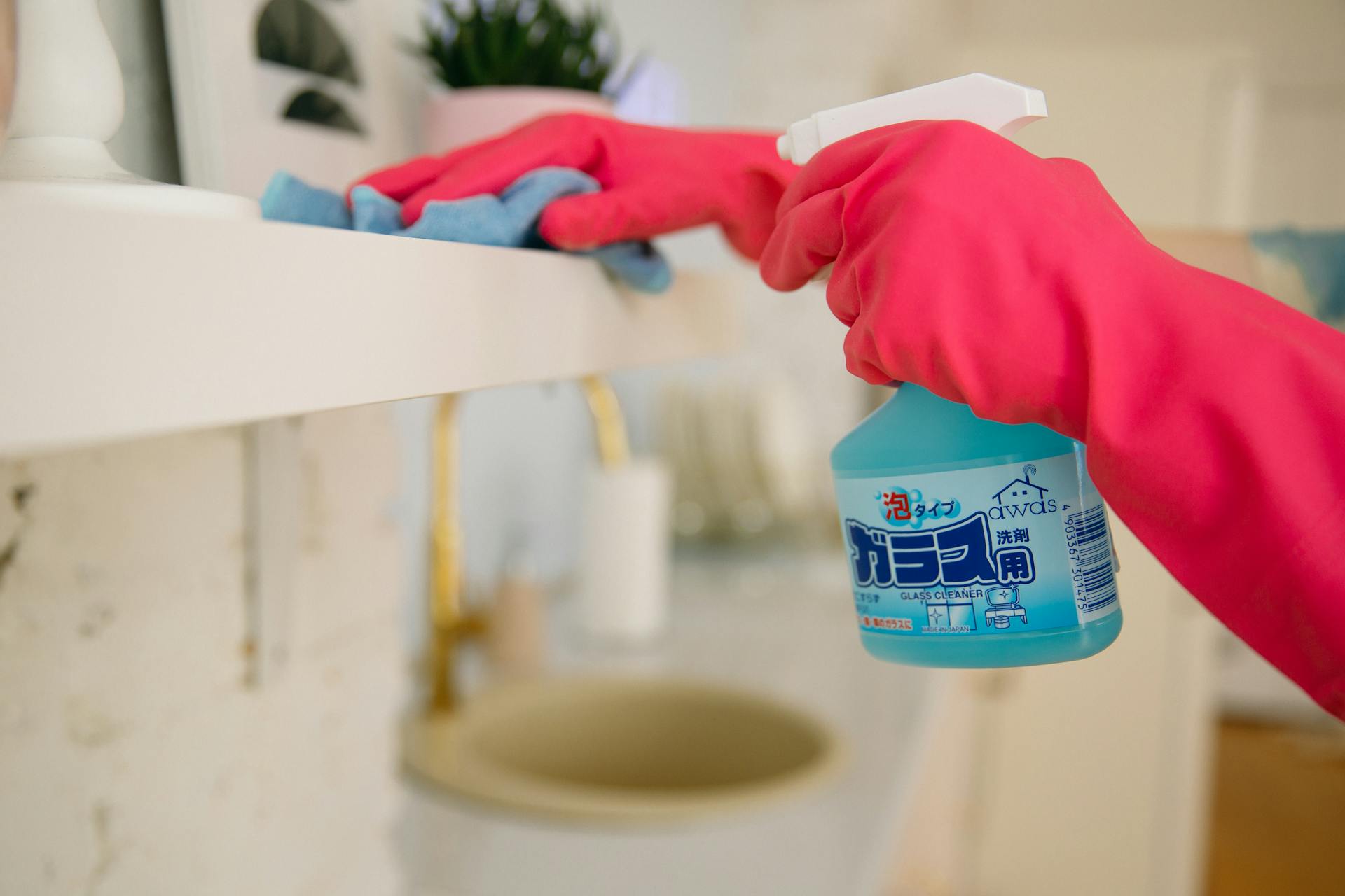 A woman using a blue spray cleaner and cloth while wearing pink gloves for home cleaning.