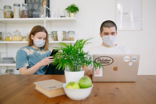 Free Two adults wearing face masks, working remotely at home with a laptop and smartphone, observing COVID-19 safety measures. Stock Photo