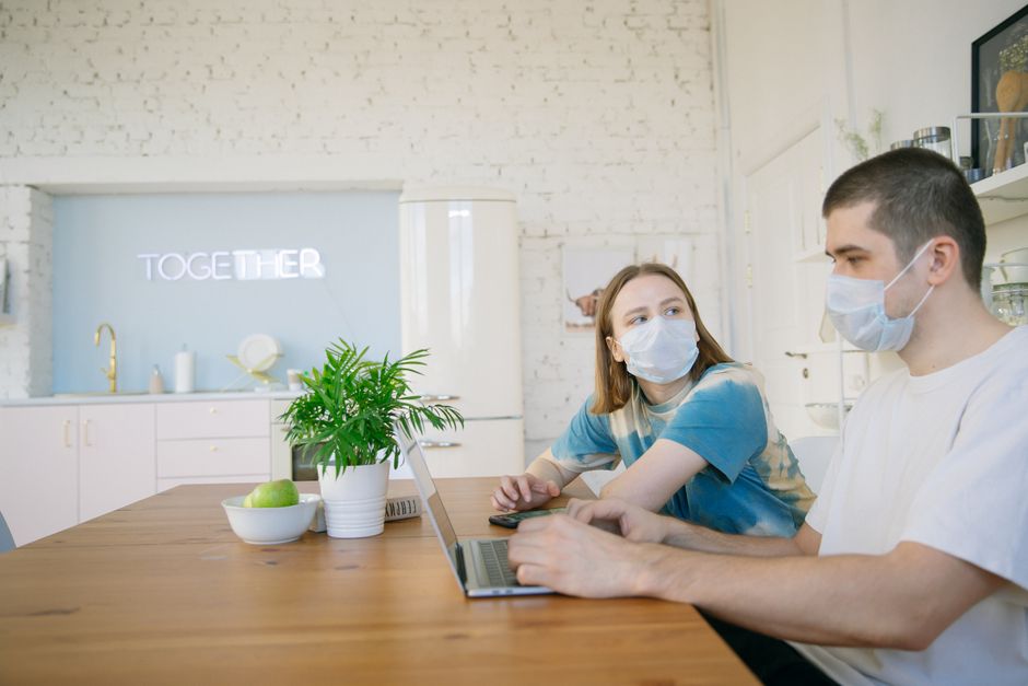 Dos jóvenes usan máscaras mientras trabajan juntos en casa, reflejando la nueva normalidad.