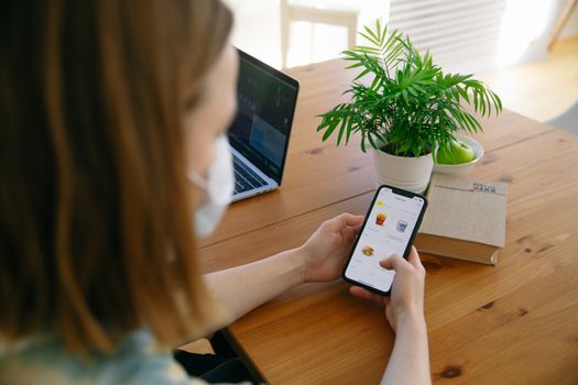 Woman using smartphone for online shopping with laptop and plant on desk indoors.
