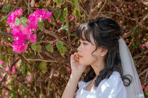 Side view of young calm ethnic female wearing veil and white wedding dress with delicate blossoming flowers on branch