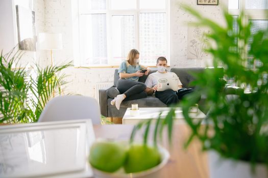 Couple sitting on sofa with masks, using electronics in bright room.