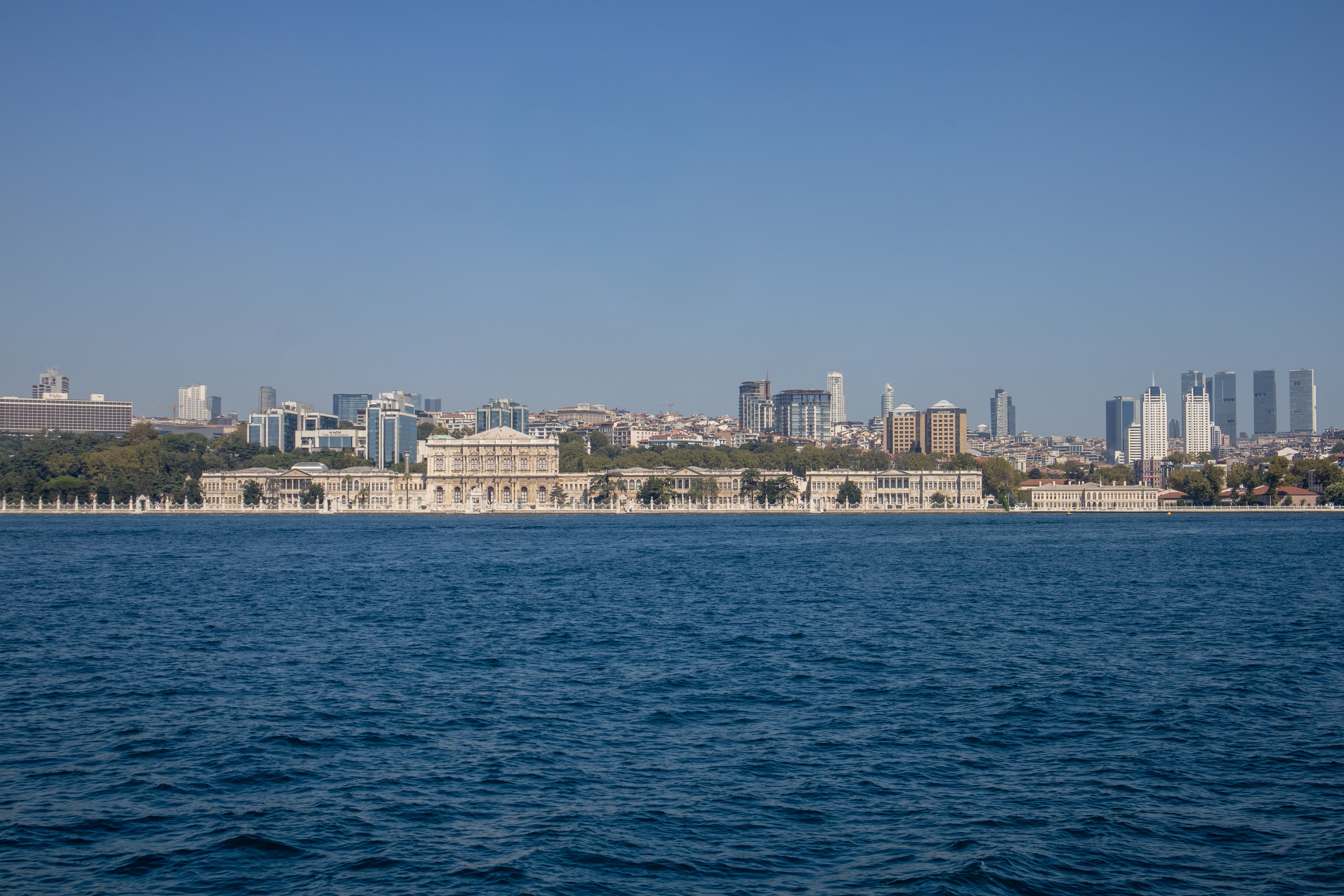A long, low cream-coloured palace on a waterfront seen across dark blue water, with city towers rising behind it