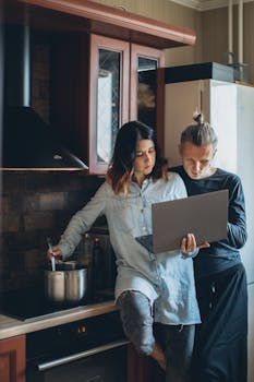 A young couple multitasking in a cozy kitchen, cooking and using a laptop. Perfect for lifestyle or remote work themes.