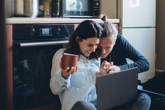 A couple sits on their kitchen floor laughing at an open laptop, one holding a terracotta mug