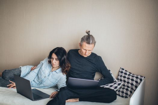 Couple working from home on laptops, sitting together on a bed.