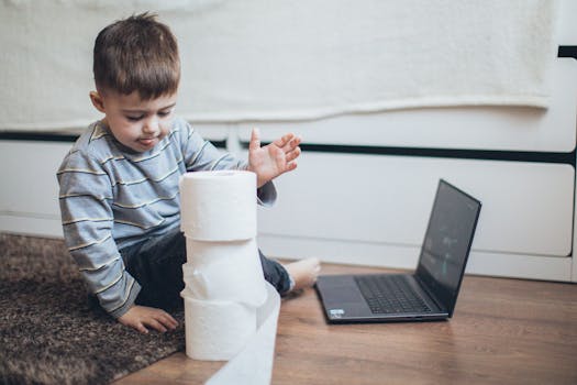 Young boy playing with tissue rolls next to a laptop indoors, reflecting fun home activities.