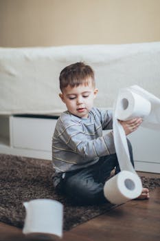 A cute child sitting on the floor indoors unraveling toilet paper with a playful expression.