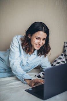 Woman smiling while working remotely from bed using a laptop.