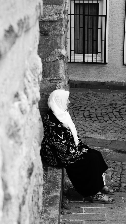 Free A woman in traditional attire sits quietly against a stone wall on a cobblestone street in İstanbul. Stock Photo
