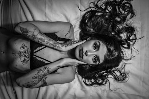 From above of black and white young tattooed sensual female in lace lingerie touching cheeks while lying on bed and looking at camera