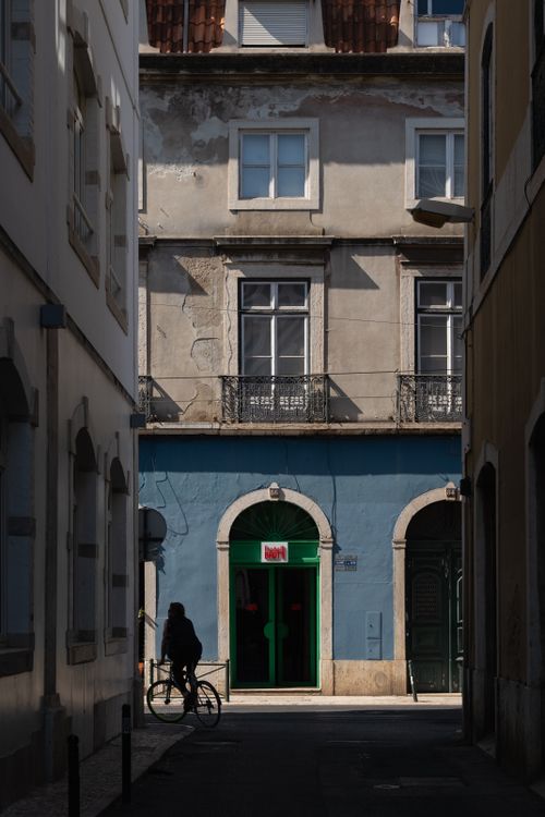 Free A shadowy alleyway in Lisbon with a person on a bicycle, revealing vibrant architecture. Stock Photo