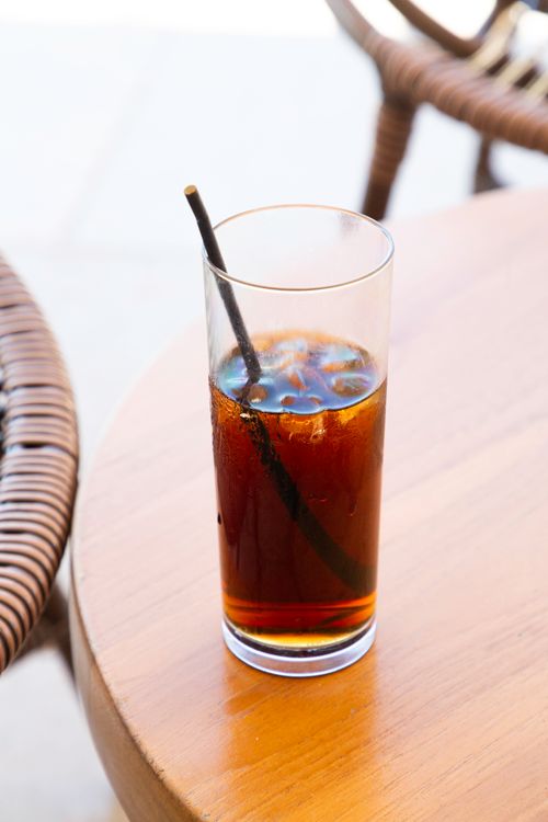 Free A tall glass of iced coffee with a straw, sitting on a wooden table in a Greek outdoor cafe. Stock Photo
