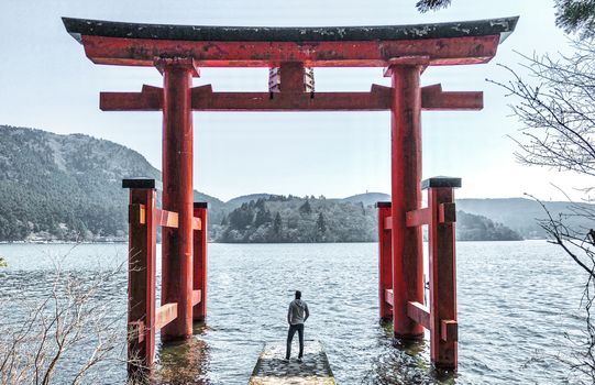 在日本的芦之湖上，一个人站在箱根神社标志性的鸟居门前，景色宁静