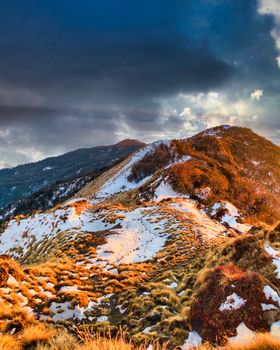 Amazing landscape of mountain range covered with dry grass and snow against sunlight shining through thick gray clouds