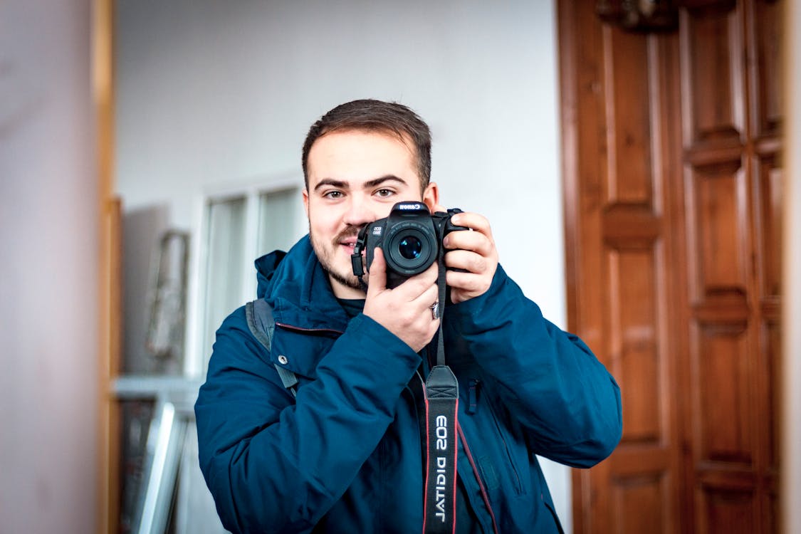 Happy young bearded male photographer in warm clothes taking mirror selfie on camera while standing against white studio interior