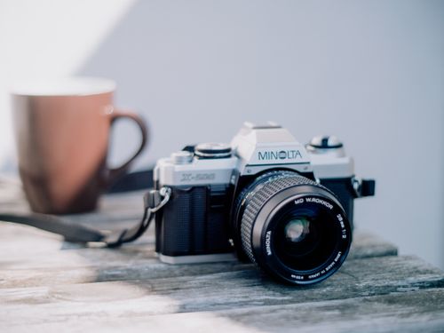 Free A vintage Minolta film camera sits on a wooden table beside a coffee mug, evoking nostalgia. Stock Photo