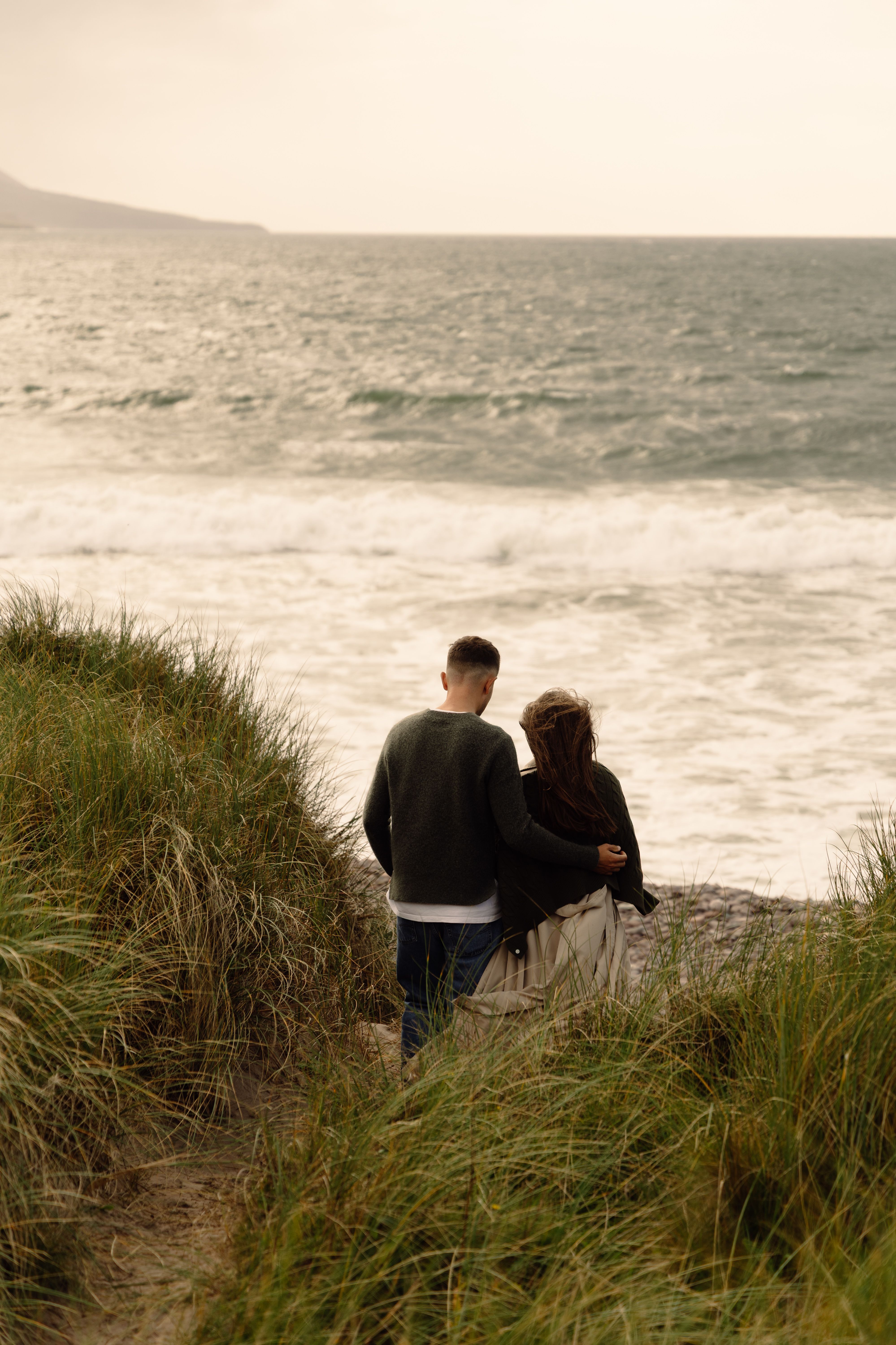 A couple walking toward the ocean through sandy dunes bordered by beach grass